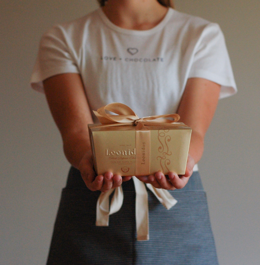 A person wearing a white t-shirt reading Love + Chocolate and a heart logo and a denim apron offers with both hands a beautiful gold box wrapped in ribbons that reads Leonidas