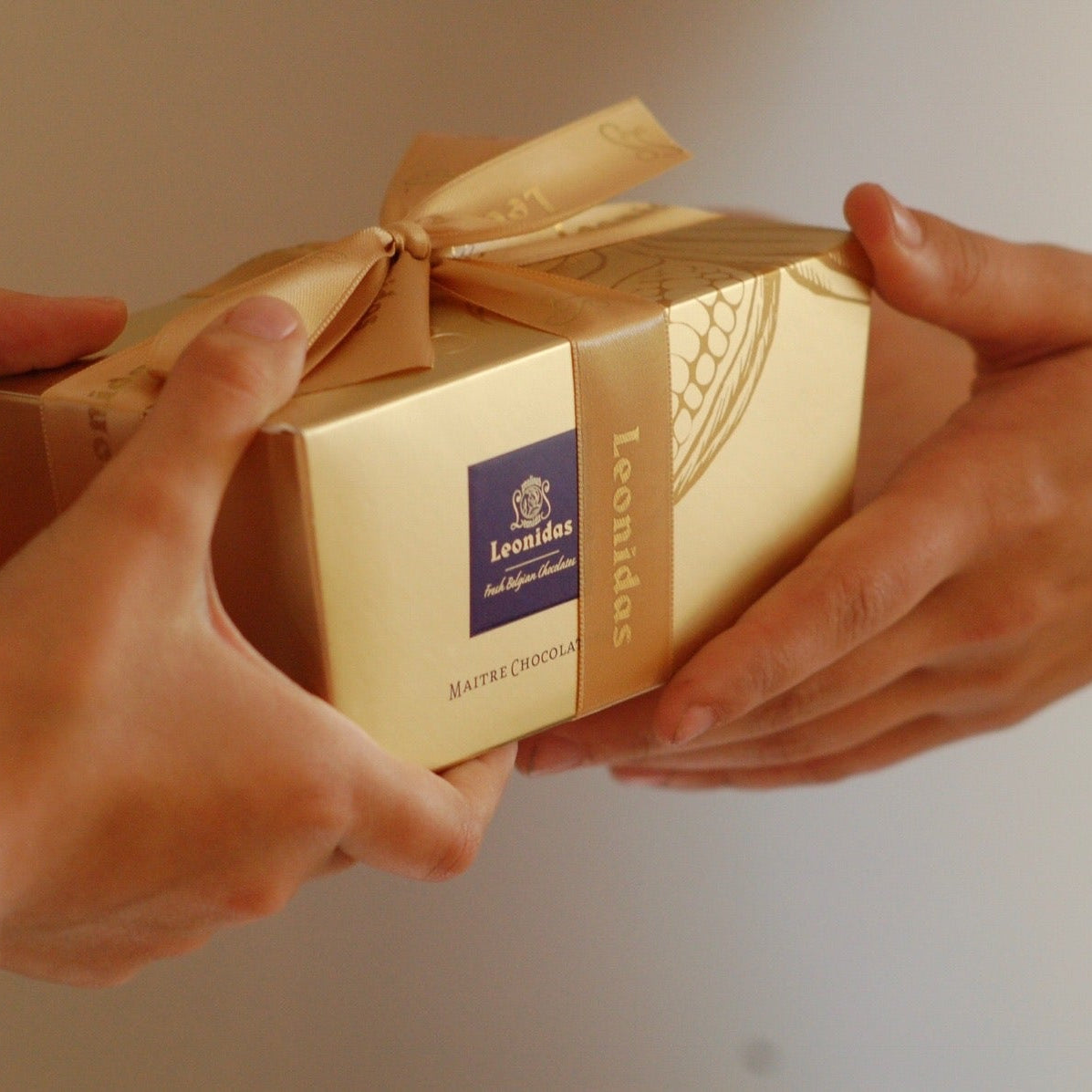 A beautiful Leonidas Gold Ballotin gift box with a gold ribbon being handed from one person to another