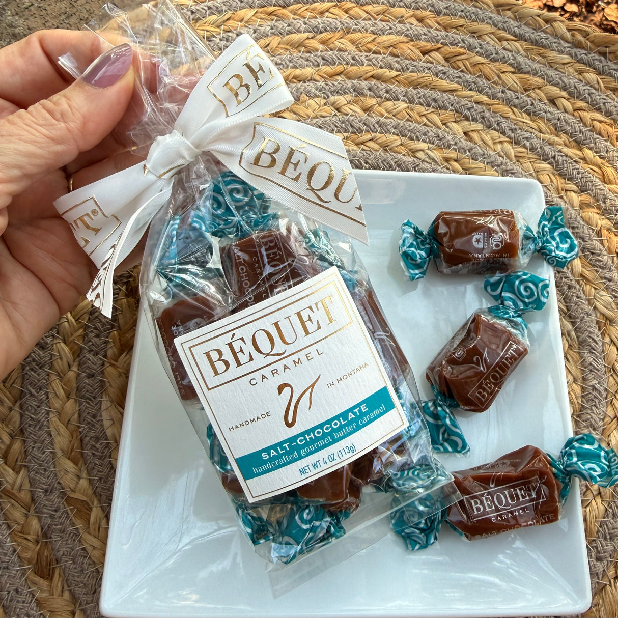 A package of Béquet Salt-Chocolate caramels with a pretty bow, on a plate with individually wrapped caramels, on a woven mat