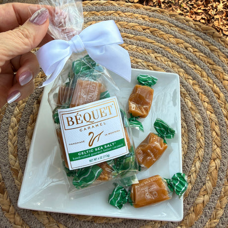 A package of Béquet Celtic sea salt caramels with a pretty bow, on a plate with individually wrapped caramels, on a woven mat