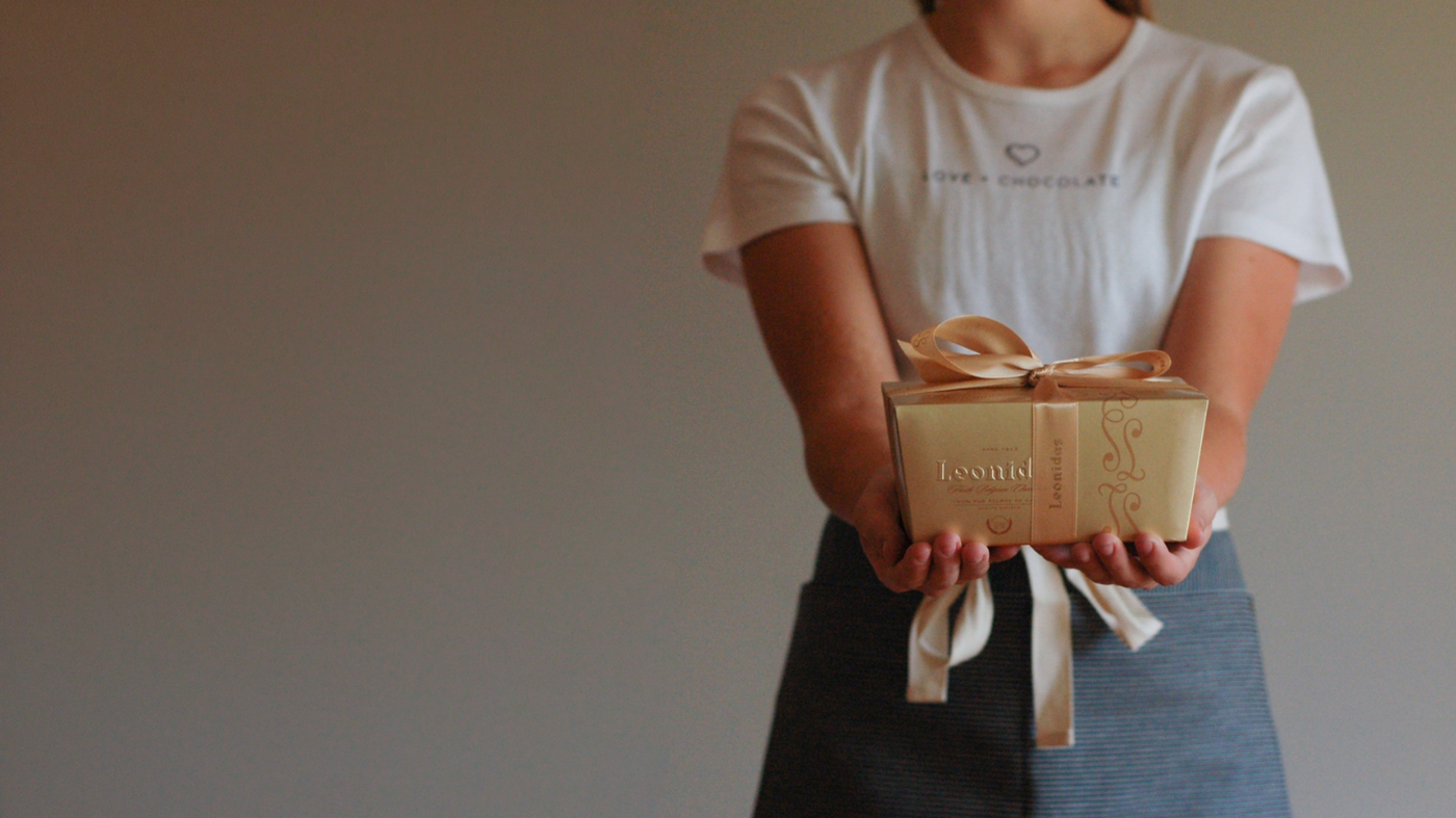 A person wearing a white t-shirt reading Love + Chocolate and a heart logo and a denim apron offers with both hands a beautiful gold box wrapped in ribbons that reads Leonidas