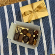 A classic Leonidas gold ballotin box with a beautiful ribbon sits next to an open box filled with an assortment of shiny dark, milk, and white chocolates, all sitting atop a pretty blue and white striped placemat