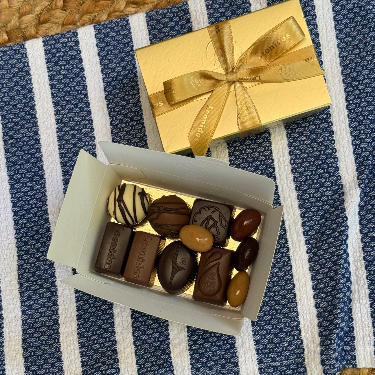 A classic Leonidas gold ballotin box with a beautiful ribbon sits next to an open box filled with an assortment of shiny dark, milk, and white chocolates, all sitting atop a pretty blue and white striped placemat