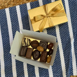 A classic Leonidas gold ballotin box with a beautiful ribbon sits next to an open box filled with an assortment of shiny dark, milk, and white chocolates, all sitting atop a pretty blue and white striped placemat