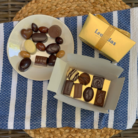 A classic Leonidas gold ballotin box with a beautiful ribbon sits next to a plate covered with a mouth-watering assortment of glossy dark, milk, and white chocolates, all sitting atop a pretty blue and white striped placemat