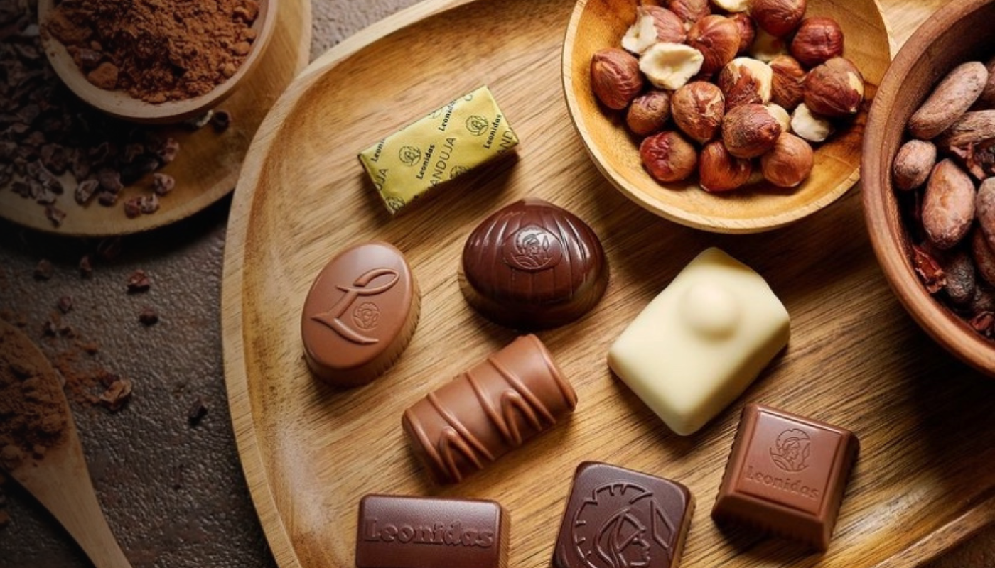 Glossy Leonidas chocolates sit on a wooden tray next to small wooden bowls filled with hazelnuts, cacao beans, cocoa nibs, and fragrant ground cocoa
