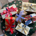 Six classic Leonidas gold ballotin boxes tied with a variety of festive ribbons sitting on a circle of green, red and white fabric decorated with mittens