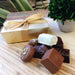 A classic Leonidas gold ballotin box with a beautiful ribbon next to a quarter-pound assortment of delicious milk, dark & white chocolates, the box and chocolates sit on a wood cutting board with houseplants behind