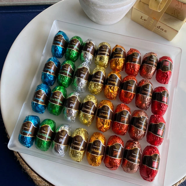 A clear tray of 32 colorful foil-wrapped delights sits atop a white table, next to a classic Leonidas gold ballotin box with a beautiful ribbon - the foils are in a rainbow of colors, each with a label that lists the liqueur within