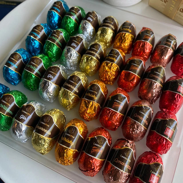 A close-up view of a clear tray of 32 colorful foil-wrapped treats - they sit atop a white table, with just a corner of a classic Leonidas gold ballotin box with a beautiful ribbon in view, and the foils are in a rainbow of colors, each with a label that lists a liqueur