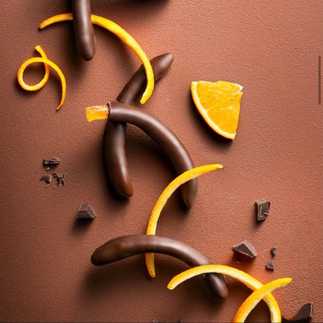 Arcs of orange rinds lay over one another, some dipped in chocolate on a light brown background with an orange segment and scattered bits of chocolate