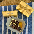 Two classic Leonidas gold ballotin boxes with beautiful ribbons sit next to an open box filled with a mouth-watering assortment of glossy dark, milk, and white chocolates, a sugar-covered fruit jelly, and a pear-shaped marzipan, all sitting atop a pretty blue and white striped placemat