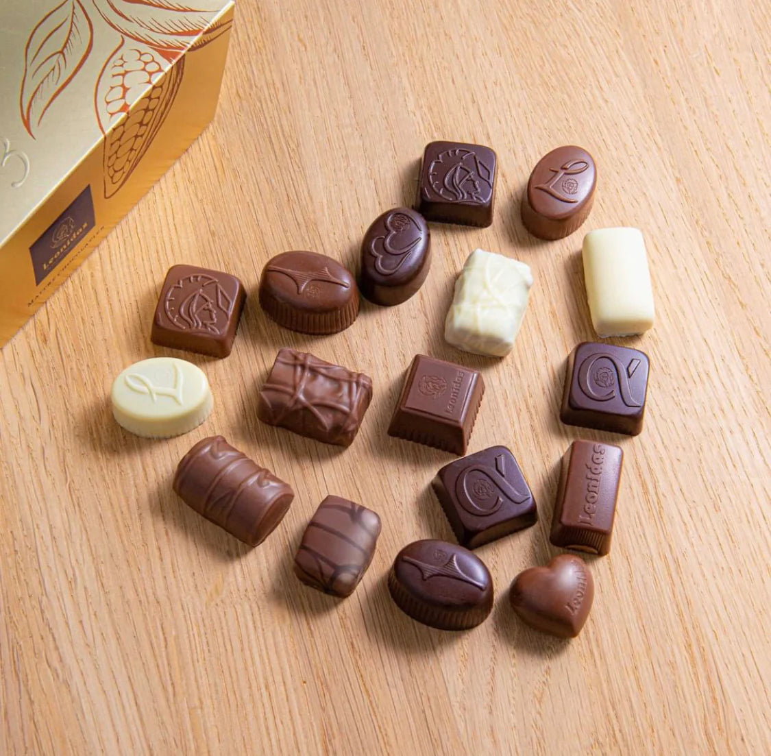 White, milk, and dark chocolates arranged in the shape of a heart on a light-wood table, next to a gold box with the Leonidas logo