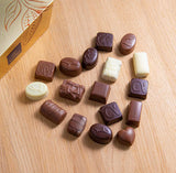 White, milk, and dark chocolates arranged in the shape of a heart on a light-wood table, next to a gold box with the Leonidas logo