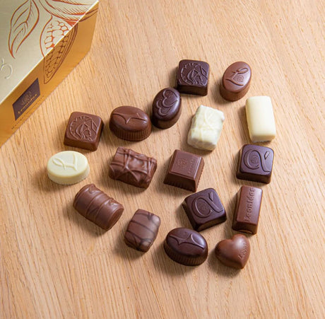 White, milk, and dark chocolates arranged in the shape of a heart on a light-wood table, next to a gold box with the Leonidas logo