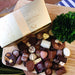 A large classic Leonidas gold ballotin box with a beautiful ribbon next to a mouth-watering assortment of glossy milk, dark & white chocolates, the box and chocolates sit on a wood table with a houseplant