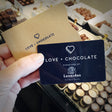 A hand holding a handsome blue card printed with 'Love + Chocolate Purveyors of Leonidas Fresh Belgian Chocolates" and a brown envelope printed with 'Love + Chocolate', above a sea of luscious chocolates in a display case