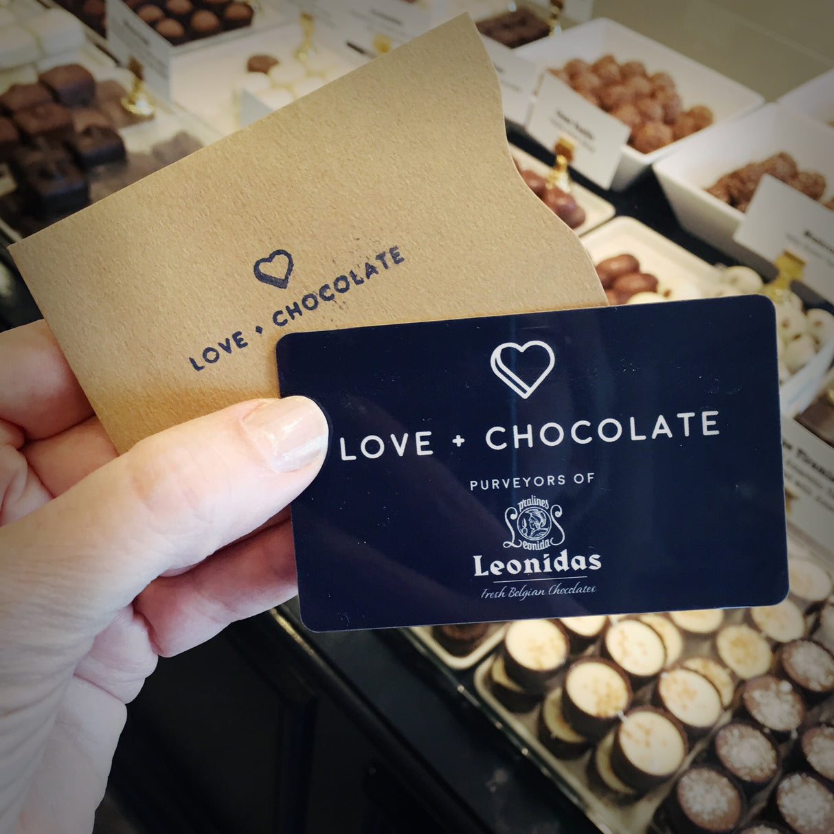 A hand holding a handsome blue card printed with 'Love + Chocolate Purveyors of Leonidas Fresh Belgian Chocolates" and a brown envelope printed with 'Love + Chocolate', above a sea of luscious chocolates in a display case